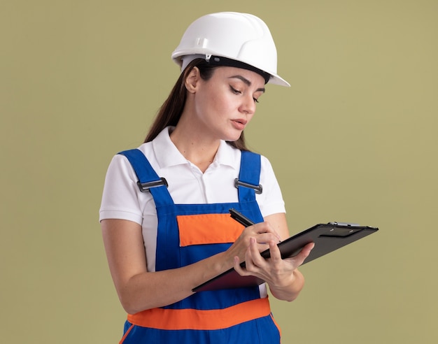 Seguros de mujer joven constructor en uniforme escribiendo algo en el portapapeles aislado en la pared verde oliva