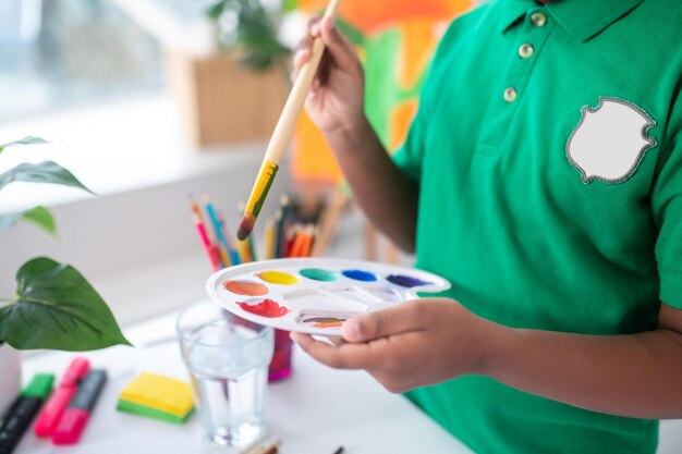 Selección de colores. Manos de un niño en edad escolar de piel oscura con camiseta verde sosteniendo un pincel sobre una paleta blanca con pinturas brillantes de pie cerca de la mesa en una habitación luminosa