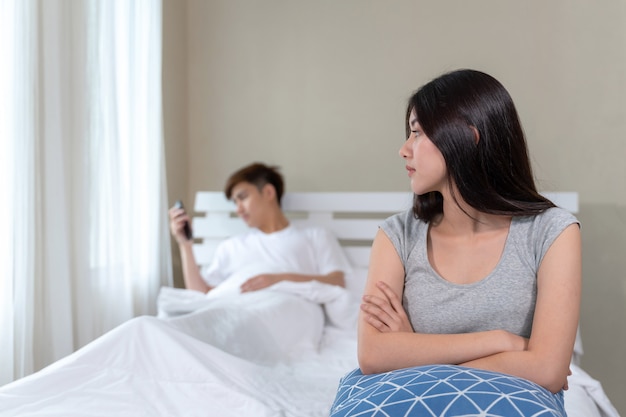 Selectiva joven esposa sentada en la cama con aburrimiento de su marido era adicto a los teléfonos inteligentes