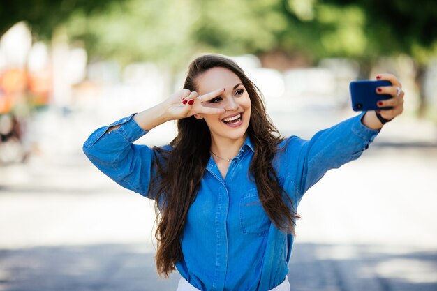 Selfie de divertida chica atractiva haciendo pucheros y mostrando el signo v en la calle