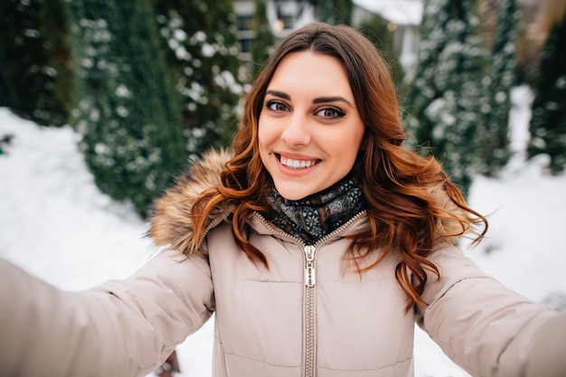 Selfie de invierno. La chica joven feliz en ropa del invierno toma el selfie en fondo nevoso del invierno
