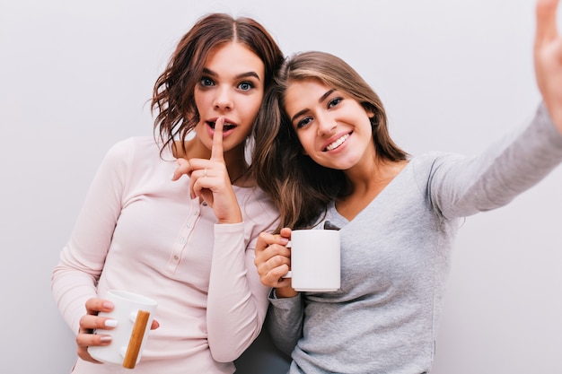 Selfie retrato de dos niñas en pijama con tazas en la pared gris. Chica con cabello rizado puso el dedo en los labios, sonriendo.