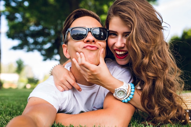 Selfie-retrato de pareja divertida tumbado en la hierba en el parque de verano. Chica con pelo largo y rizado, labios rojos y chico joven con gafas de sol imitando a la cámara.