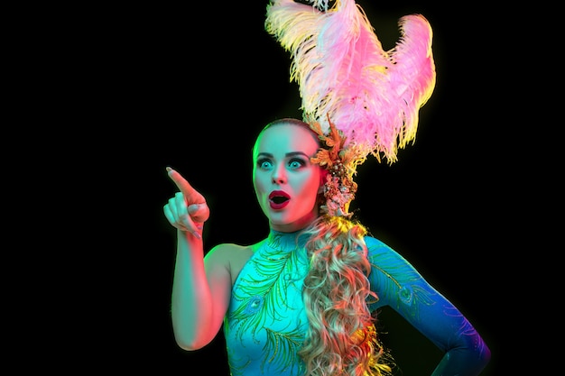 Señalando. Hermosa mujer joven en carnaval, elegante disfraz de mascarada con plumas en pared negra en luz de neón. Copyspace para anuncio. Celebración de fiestas, baile, moda. Tiempo festivo, fiesta.