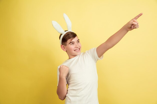 Señalando a un lado. Chico caucásico como un conejito de Pascua sobre fondo amarillo de estudio. Felices saludos de pascua. Hermoso modelo masculino. Concepto de emociones humanas, expresión facial, vacaciones. Copyspace.