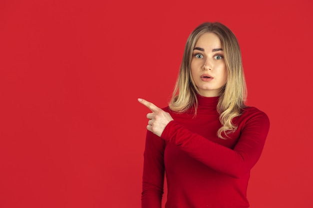 Señalando. Retrato monocromo de joven mujer rubia caucásica aislada en la pared roja del estudio.