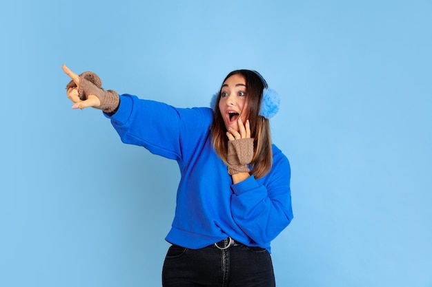 Señalando. Retrato de mujer caucásica sobre fondo azul de estudio. Modelo de mujer hermosa en ropa de abrigo. Concepto de emociones humanas, expresión facial, ventas, publicidad. Estado de ánimo de invierno, Navidad, vacaciones.