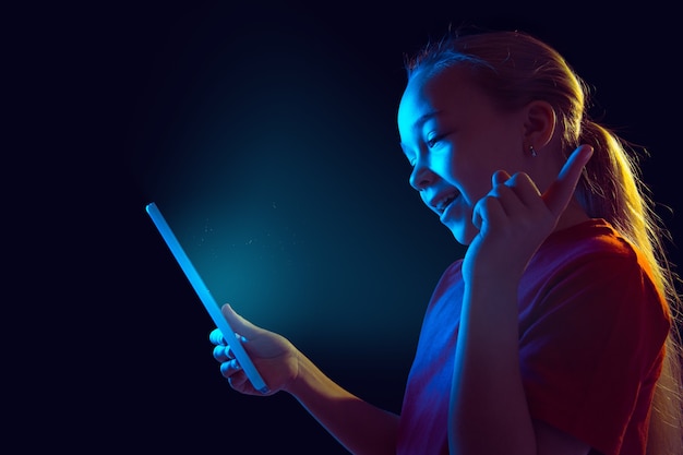 Señalando, riendo. Retrato de niña caucásica en la pared oscura en luz de neón. Modelo femenino hermoso que usa la tableta. Concepto de emociones humanas, expresión facial, ventas, publicidad, tecnología, gadgets.