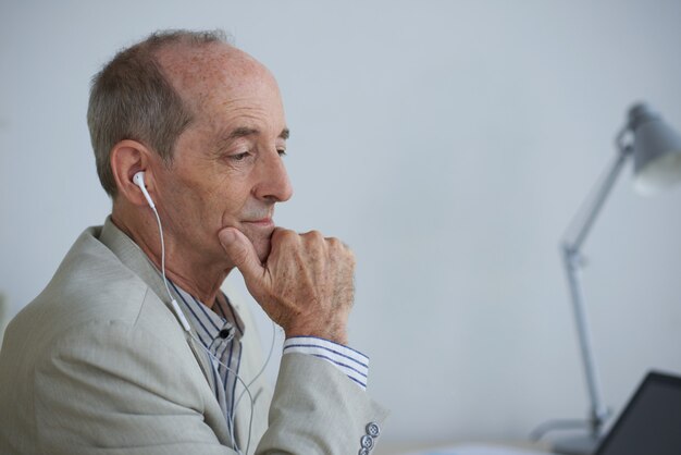 Senior empresario caucásico sentado en la oficina con auriculares y mirando portátil