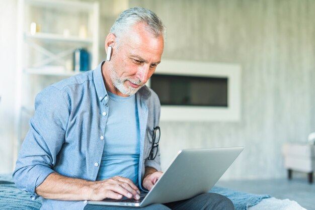 Senior hombre con bluetooth inalámbrico sentado en la cama usando la computadora portátil