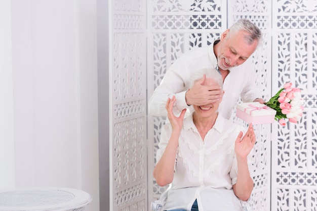 Senior hombre cubriendo los ojos de su esposa con una mano y dándole un regalo