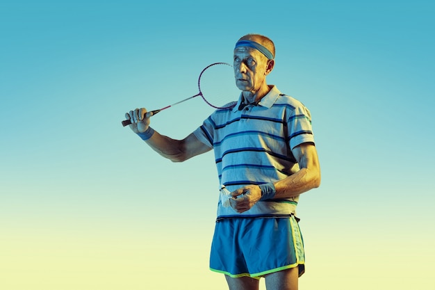 Senior hombre vestido con ropa deportiva jugando bádminton sobre fondo degradado, luz de neón.