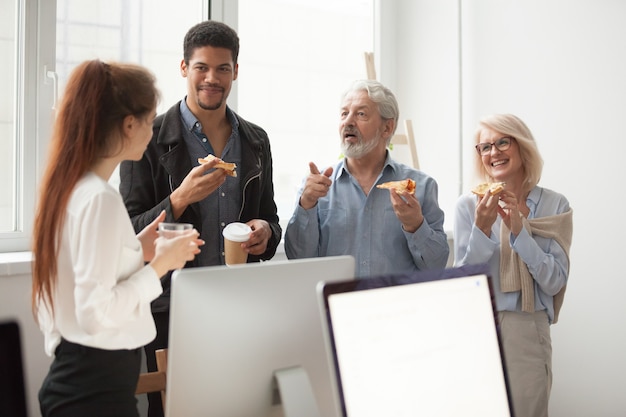 Senior y jóvenes colegas hablando mientras comen pizza en la oficina