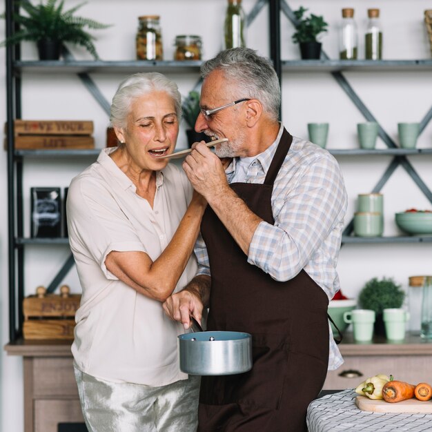 Senior mujer degustación de comida preparada por su esposo en la cocina