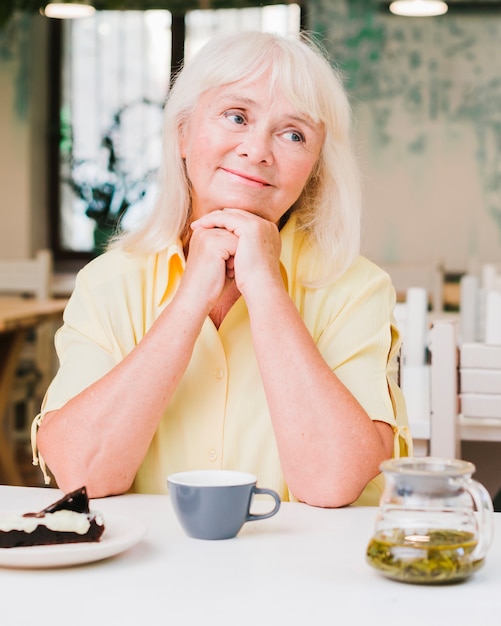 Senior mujer sentada en la cocina con taza y pastel