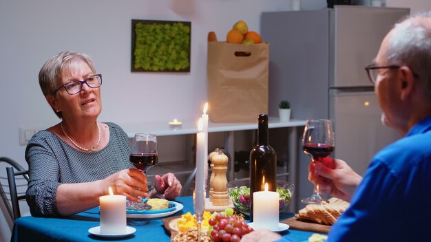 Senior pareja relajada cenando y bebiendo vasos de vino tinto juntos en la cocina de casa. Ancianos, ancianos jubilados disfrutando de la comida, celebrando su aniversario en el comedor.