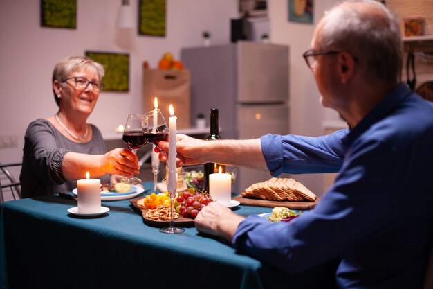 Senior pareja sosteniendo copas de vino durante la celebración de la relación en la cocina por la noche. Pareja de ancianos sentados a la mesa en el comedor, hablando, disfrutando de la comida,