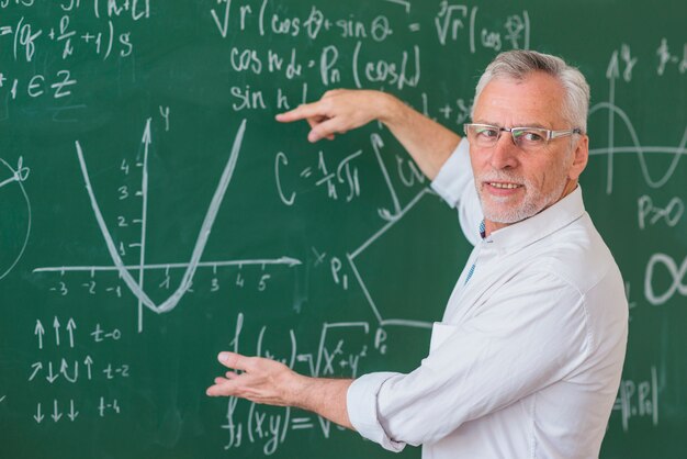 Senior profesor en gafas explicando ejemplo de matemáticas en pizarra verde