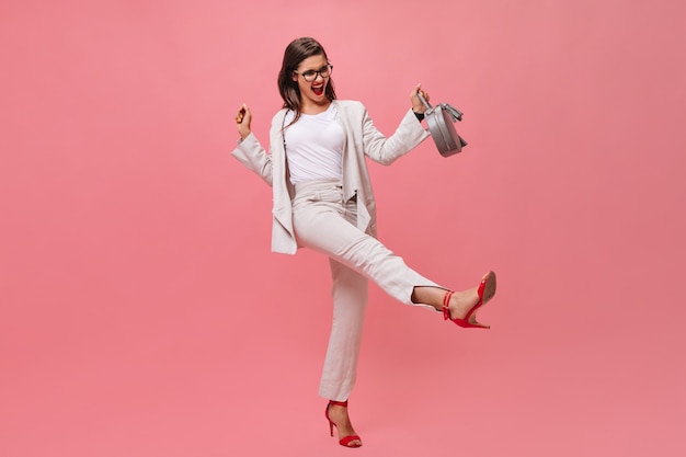 Señora activa en traje posando con bolso sobre fondo rosa. Mujer joven emocional que se divierte en la cámara en tacones rojos.