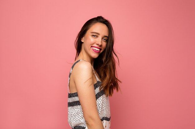 Señora adorable feliz con el pelo largo y ondulado en vestido de verano posando sobre fondo rosa