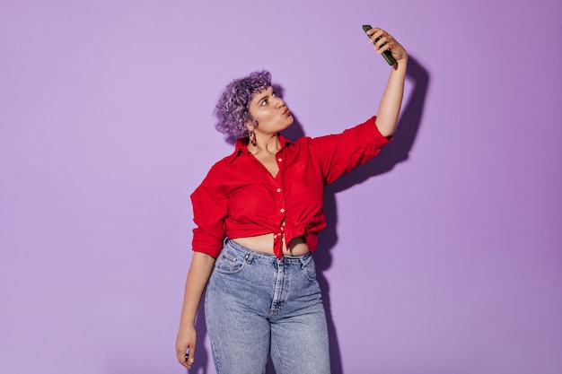 Foto gratuita señora adulta brillante en ropa inusual elegante hace foto en lila. mujer rizada de pelo corto en camisa roja toma selfie.