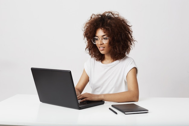 Señora africana del asunto que sonríe trabajando en la computadora portátil sobre la pared blanca.