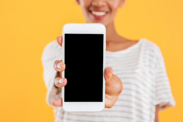 Señora africana feliz que muestra el teléfono con la pantalla en blanco aislada