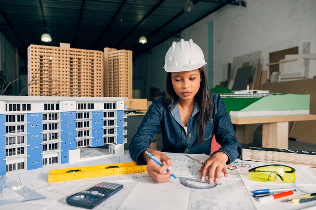 Señora afroamericana en casco de seguridad trabajando cerca de modelo de edificio