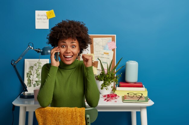 Señora afroamericana positiva aprieta el puño mientras habla por teléfono inteligente, se siente muy feliz, sonríe ampliamente, trabaja en el informe financiero en casa