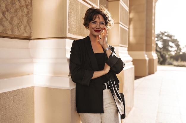 Señora alegre en chaqueta y pantalón blanco sonriendo afuera. Mujer encantadora con labios brillantes en anteojos al aire libre.