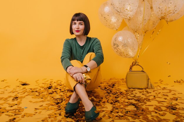 Señora alegre con el pelo corto posando con expresión de la cara interesada. Retrato interior de la hermosa cumpleañera escalofriante con confeti y globos.