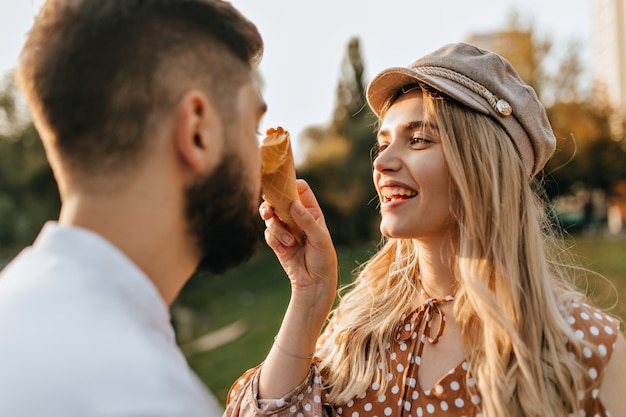 Señora alegre con sombrero elegante y top de lunares se ríe, manchando la nariz de su marido con helado de melón.