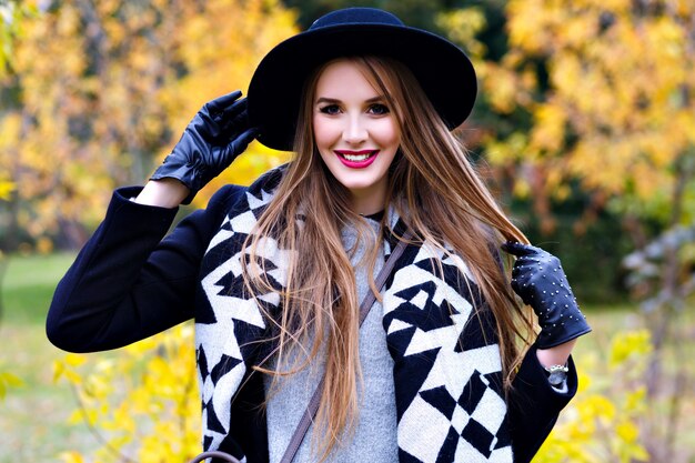 Señora alegre con sombrero negro y guantes jugando con el pelo largo con bosque de fondo. Hermosa chica con abrigo y elegante bufanda sonriendo durante la caminata en el parque de otoño.
