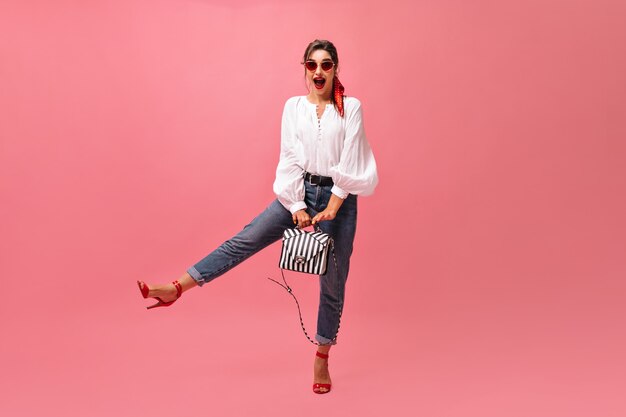 Señora en anteojos redondos posa con bolso sobre fondo rosa. Mujer joven positiva en gafas de sol rojas y labios brillantes se regocija en la cámara.