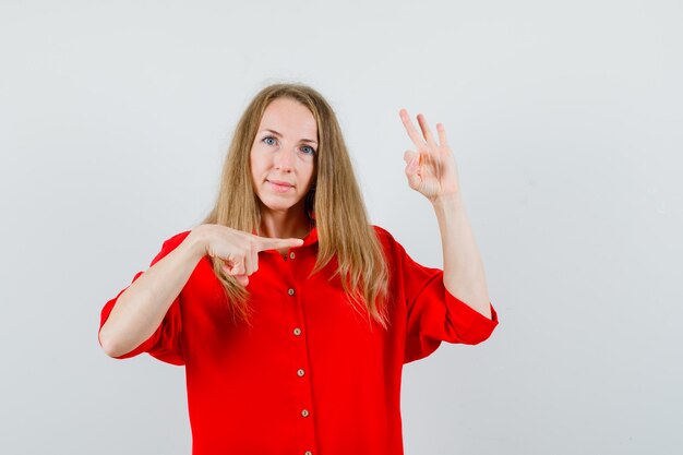 Señora apuntando a un lado, mostrando el signo de ok en camisa roja y mirando confiada.