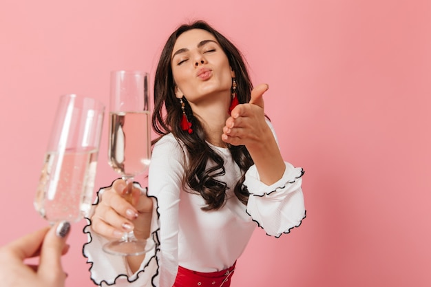 Foto gratuita señora con aretes rojos y blusa blanca envía beso al aire y tintinea vasos de vino espumoso sobre fondo aislado.