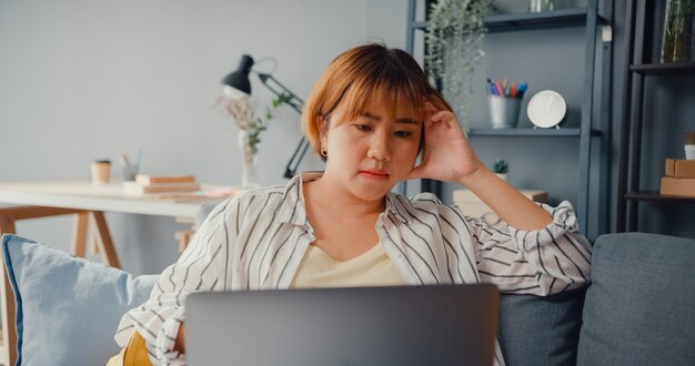 Señora asiática independiente siente dolor de cabeza mientras está sentada en el sofá con la computadora portátil en línea, aprende en la sala de estar de la casa