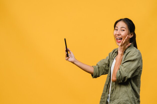 Señora asiática joven sorprendida que usa el teléfono móvil con expresión positiva, sonríe ampliamente, vestida con ropa casual y está aislada en la pared amarilla. Feliz adorable mujer alegre se regocija con el éxito.