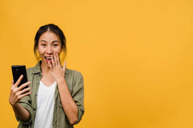 Señora asiática joven sorprendida que usa el teléfono móvil con expresión positiva, sonríe ampliamente, vestida con ropa casual y está aislada en la pared amarilla. Feliz adorable mujer alegre se regocija con el éxito.