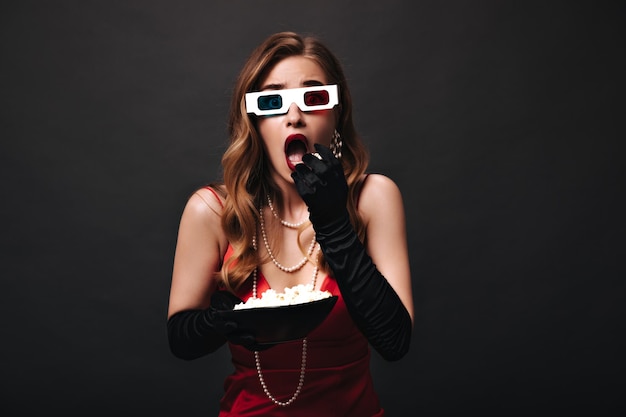 Señora asombrada en gafas 3D comiendo palomitas de maíz sobre fondo negro