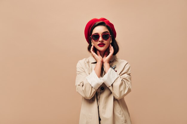 Señora atractiva en gafas de sol y poses de boina roja sobre fondo beige. Chica maravillosa en abrigo ligero de otoño y sombrero brillante mira a la cámara.