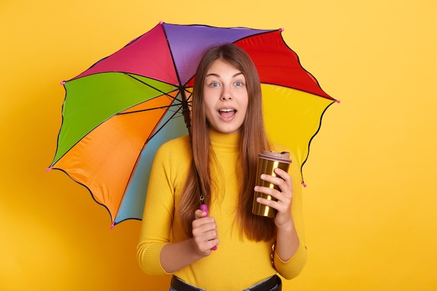 Señora atractiva joven con expresión facial asombrada posando con paraguas multicolor y café para llevar, se encuentra con la boca abierta