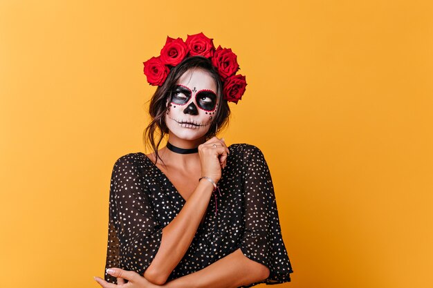 Señora atractiva con maquillaje de Halloween mira pensativamente. Retrato de niña con flores rojas en el pelo posando sobre fondo naranja.