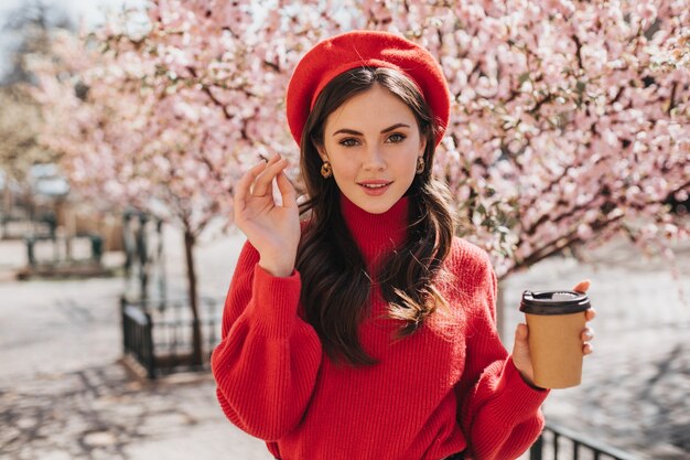 Señora atractiva en suéter rojo camina por la avenida con sakura y bebe café. Bella mujer en boina sonriendo y disfrutando de un té fuera