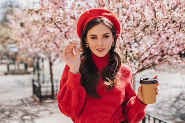 Señora atractiva en suéter rojo camina por la avenida con sakura y bebe café. Bella mujer en boina sonriendo y disfrutando de un té fuera