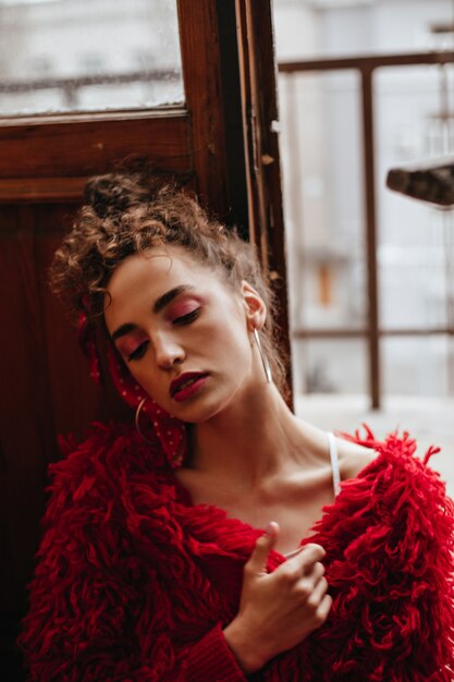 Señora atractiva en suéter rojo posando junto a la ventana