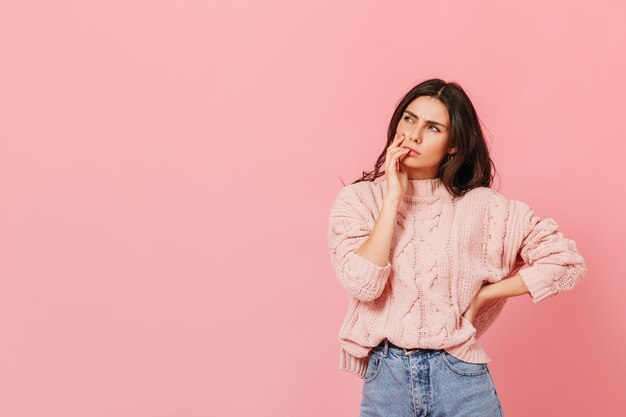 Señora atractiva en traje brillante está pensando en una nueva idea. Mujer en suéter posando pensativamente sobre fondo rosa.