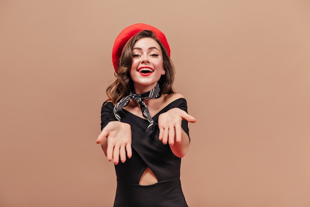 Señora atractiva en vestido negro y sombrero rojo está sonriendo y mostrando sus palmas.