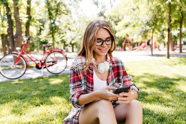 Señora blanca dichosa usando el teléfono en el parque. Chica europea alegre en gafas casuales sentado en la hierba y mensaje de texto.