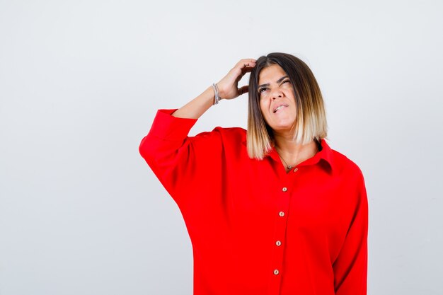 Señora bonita en blusa roja rascándose la cabeza, mirando hacia arriba y mirando olvidadizo, vista frontal.